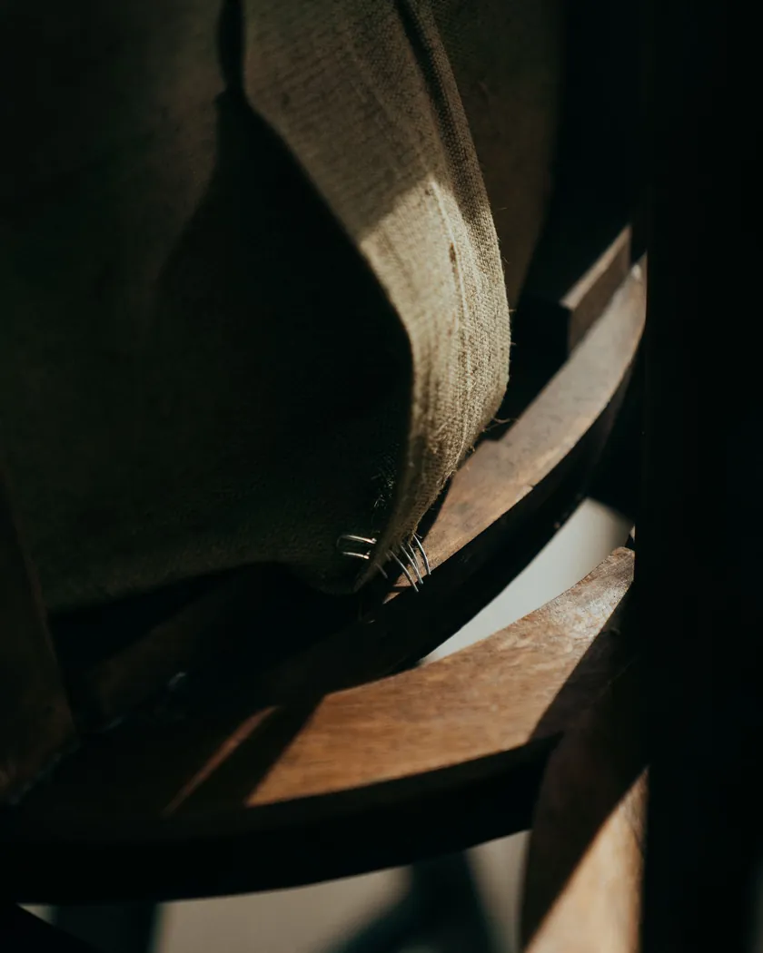 Primer plano de una arpillera grapada sobre el armazón curvado de madera de un asiento, con las grapas a la vista