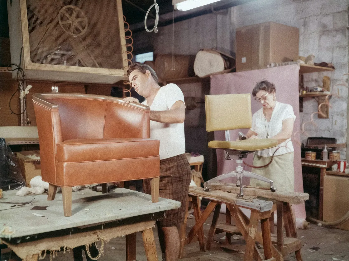 Interior de un taller de tapicería con una butaca forrada de piel sobre un caballete y otra silla en proceso de trabajo